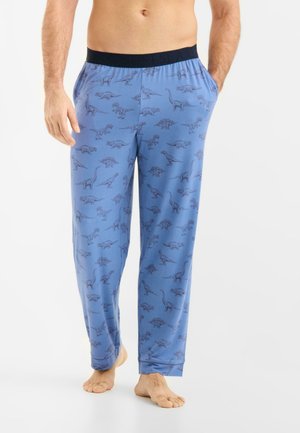 Mies, joka pukeutunut sinisiin pyjamanhousuihin, joissa on tummia dinosauruskuvioita, seisoo paljain jaloin, toinen käsi taskussa, yksinkertaista valkoista taustaa vasten.