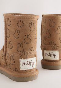 Braune Veloursleder-Stiefel mit einem sich wiederholenden Miffy-Hasenmuster, weichem Innenfutter und einem gebrandeten weißen Etikett an der Seite mit braunen Textakzenten.