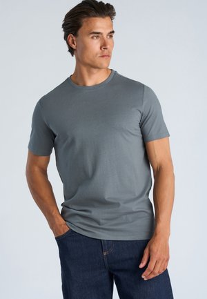 Man met kort donker haar, gekleed in een grijs T-shirt met korte mouwen en donkerblauwe spijkerbroek, staand met één hand in de zak en kijkend naar rechts.