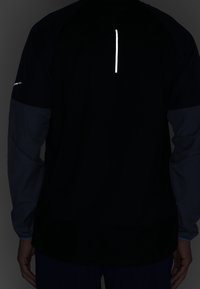 Schwarze Jacke mit langen Ärmeln und grauen Ärmeln, ausgestattet mit einem reflektierenden vertikalen Streifen auf dem Rücken und einer lässigen Passform. Weiche, glatte Textur.