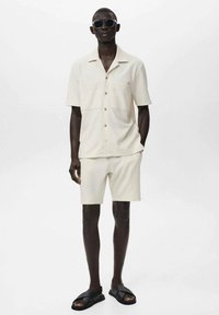 Lys beige kortærmet skjorte med to brystlommer, parret med matchende shorts. Båret med sorte solbriller og sandaler. Glat tekstur.