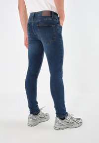 Vaqueros de mezclilla en azul oscuro, de corte slim, con bolsillos traseros y detalles de costura. Llevados con zapatillas deportivas plateadas.