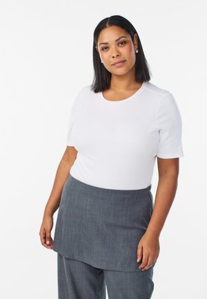 Kvinde med skulderlangt mørkt hår iført en hvid kortærmet top og gråt lagdelt nederdel og bukser foran en hvid baggrund.