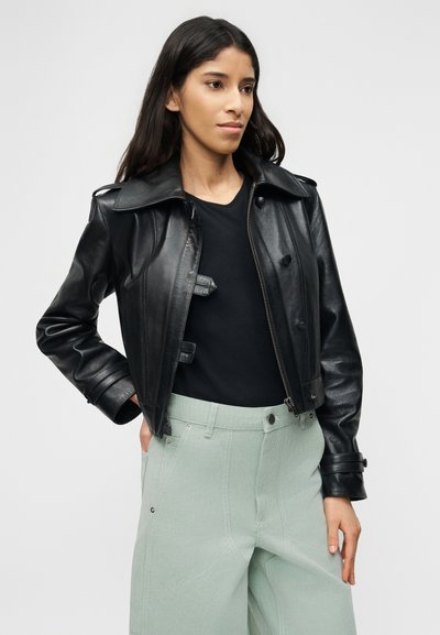 Γυναίκα με μακριά σκούρα μαλλιά, ντυμένη με μαύρο δερμάτινο μπουφάν πάνω από μαύρο τοπ και ανοιχτό πράσινο παντελόνι ψηλόμεσο, στέκεται με το ένα χέρι στην τσέπη.