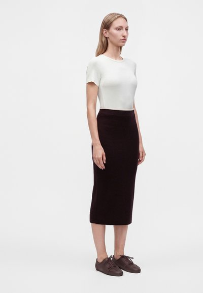 Femme debout, portant un haut blanc à manches courtes, une jupe midi noire et des chaussures marron à lacets, sur un fond clair uni.