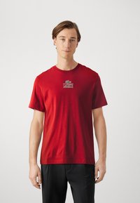 Červené bavlnené tričko s okrúhlym výstrihom a krátkymi rukávmi, s logom Lacoste v kontrastných farbách na hrudi.