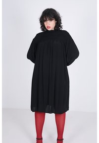 Zwarte oversized jurk met een geborduurd lijfje, driekwart mouwen en een losse pasvorm. Gepaard met rode panty's en zwarte hakken.