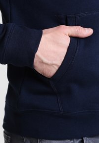 Sudadera azul marino con un acabado suave y texturizado. Cuenta con un bolsillo frontal y puños acanalados. Primer plano de una mano descansando en el bolsillo.