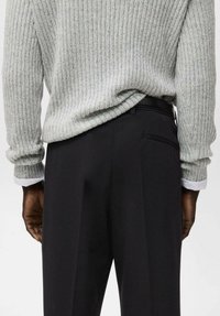 Uomo che indossa un maglione grigio chiaro a coste infilato in pantaloni neri su misura, mostrato dalla vita a metà coscia, in piedi dritto.