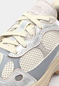 Sneaker med en blandning av material; ljusgrå mesh med silver- och ljusblå detaljer. Har tunna snören, rundad form och texturerade detaljer.