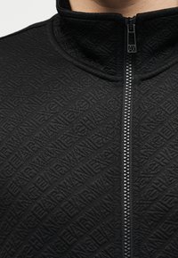 Κοντινή λήψη μαύρου υφασμάτινου μπουφάν με πλήρη φερμουάρ και ανάγλυφο επαναλαμβανόμενο μοτίβο κειμένου "ARMANI EXCHANGE" στο ύφασμα.