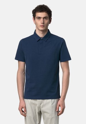 Giovane uomo in piedi, indossa una polo blu navy a maniche corte e pantaloni beige chiaro, rivolto in avanti su uno sfondo semplice.