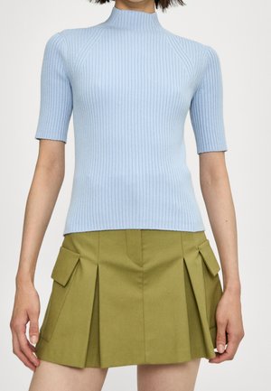 Mujer vestida con un suéter de cuello alto de manga corta acanalado color azul claro y una minifalda plisada verde oliva con bolsillos laterales con solapa.