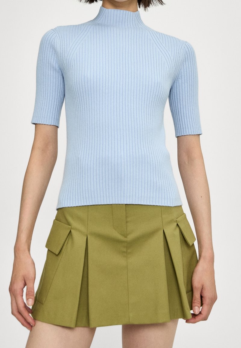 Femme portant un pull à col roulé à manches courtes en côte bleu clair et une mini-jupe plissée vert olive avec des poches à rabat sur les côtés.