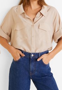 Camisa corta de mangas beige claro con cuello, dos bolsillos en el pecho y cierre de botones, combinada con jeans de mezclilla azul de cintura alta.