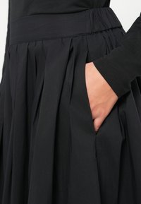 Melnā plīšu svārki ar platām jostām un sānu kabatām, izgatavoti no viegla auduma ar gludu tekstūru.