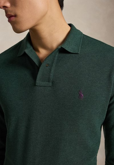 Man draagt een donker groen poloshirt met lange mouwen en een klein paars geborduurd logo op de linkerborst.
