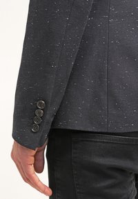 Ciemny blezer z tkaniny o teksturowanej powierzchni z białymi kropkami. Na mankiecie widoczne jest pięć brązowych guzików, co podkreśla dopasowany krój.