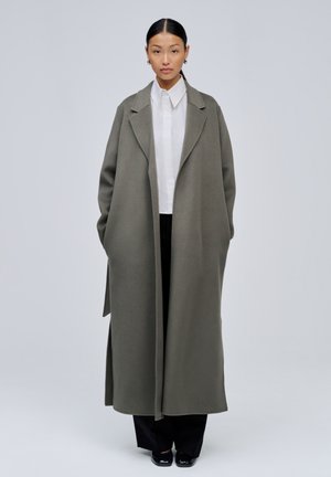 Femme portant un long manteau gris oversize par-dessus une chemise blanche et un pantalon noir, debout les mains dans les poches du manteau, sur un fond uni.