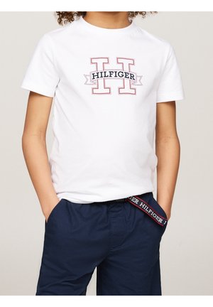 Barn iført hvid T-shirt med korte ærmer med stort omridset "H" og "HILFIGER" tekst, kombineret med mørkeblå shorts og hænderne i lommerne.