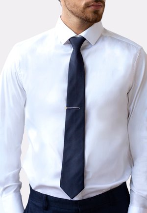 Cravate en soie bleue marine avec une extrémité pointue, maintenue par une pince à cravate dorée, portée sur une chemise blanche à col classique.