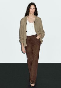 Bruna mocka byxor i kombination med en beige texturerad cardigan och en vit topp. Modellen bär ett enkelt halsband och sandaler med öppen tå.