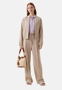 Beige Cropped-Jacke, hellviolettes Poloshirt, beige weitem Bein-Hose und Sandalen; hält eine geflochtene Tasche mit braunen Akzenten. Glatte Texturen.