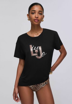 Donna con orecchini e braccialetti d'oro che indossa una maglietta nera con logo e slip bikini con stampa leopardata su sfondo semplice.