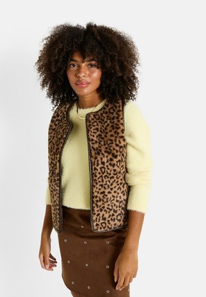 Chaleco de estampado de leopardo con una textura suave, ribete marrón y sin cierre frontal, llevado sobre un suéter de lana amarillo y una falda marrón con acentos de metal.