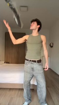 Νέος άνδρας με casual ρούχα πετάει ένα αθλητικό παπούτσι σε ένα υπνοδωμάτιο με κρεβάτι, ντουλάπα και φωτεινούς τοίχους.