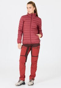 Doudoune rouge avec des coutures verticales et un col montant, accompagnée de pantalons techniques rouges et bruns dotés de poches zippées et de renforts aux genoux.
