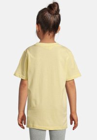 Enfant aux cheveux bruns en chignon, portant un t-shirt jaune uni à manches courtes et un legging gris, de dos sur un fond blanc.