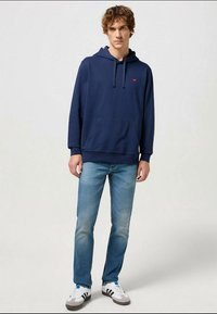 Sweatshirt de capuz azul-marinho com cordão, bolso canguru, punhos canelados, combinado com jeans de lavagem clara e ténis brancos.