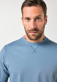 Niebieska bluza wykonana z miękkiego materiału, z okrągłym dekoltem i widocznym przeszyciem przy szyi, noszona przez mężczyznę z brodą.