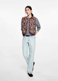 Chaleco de lana mezclada en base marrón, bordado floral en crema y verde, combinado con una blusa de manga larga gris y vaqueros de pierna ancha en un lavado claro.