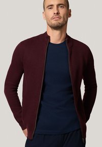 Mężczyzna w ciemno-burgundowej teksturowanej kurtce na zamek, noszący granatowy t-shirt, stojący z rękami w kieszeniach na jednolitym tle.