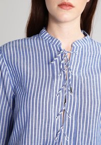 Chemise rayée bleu et blanc en tissu léger, dotée d'un col mao, d'un devant lacé et d'une finition texturée.