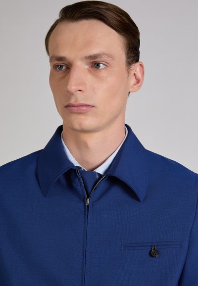 Man with light skin and brown hair wearing a blue zip-up jacket with a buttoned chest pocket, looking slightly to the left.