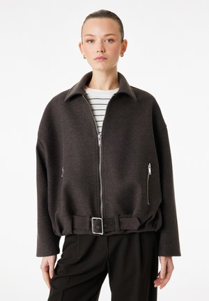 Donna che indossa una giacca con cerniera grigio scuro con colletto, tasche laterali con cerniera, fibbia e una camicia a righe sotto, in piedi contro uno sfondo bianco.