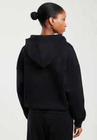 Naine, kellel on juuksed pundis ja kes kannab kuldsete rõngaskõrvarõngastega ning lahtist musta kapuutsiga dressipluusi, nähtud tagantpoolt ühetaolise heleda tausta vastu.