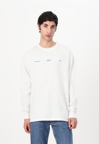 White long-sleeve t-shirt made of cotton, featuring a round neckline and purple printed logo text across the front. Worn with blue jeans.