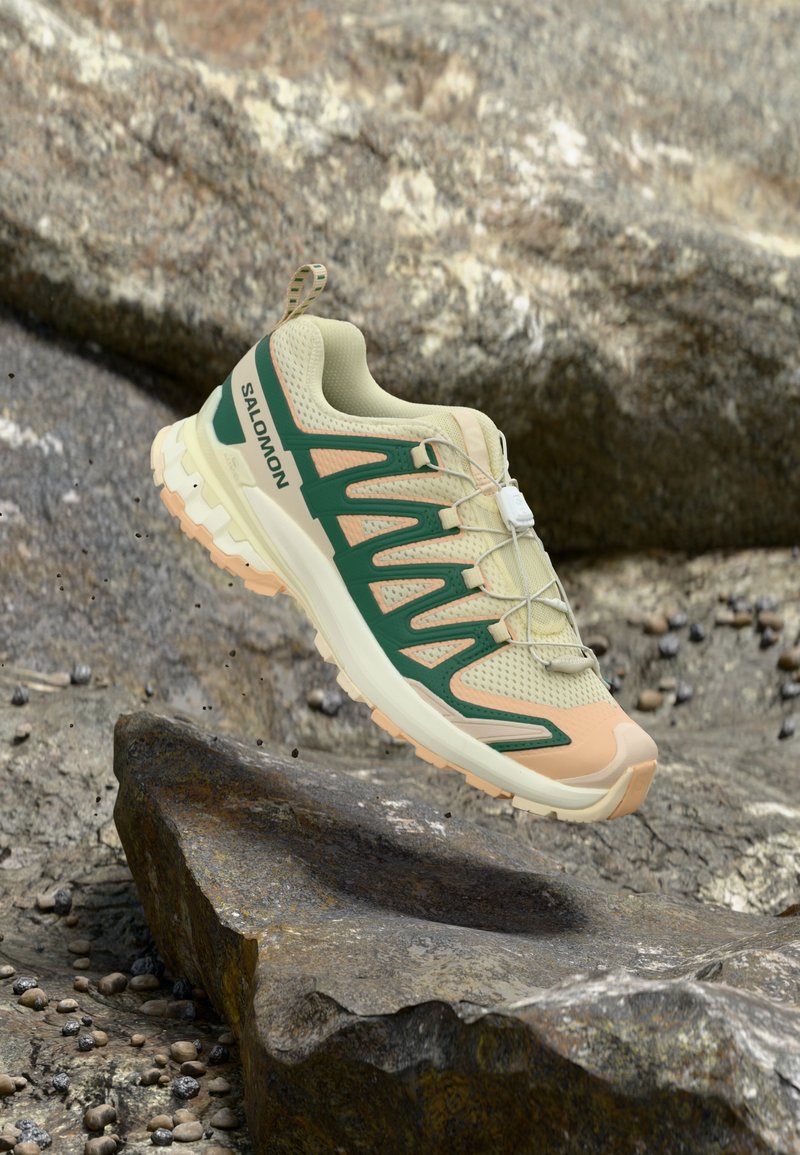 Ελαφρύ, μπεζ αθλητικό παπούτσι με πράσινες λεπτομέρειες, επάνω μέρος από δίχτυ, μοναδικός σχεδιασμός κύματος και υφή στην σόλα, τοποθετημένο σε πετρώδη επιφάνεια.