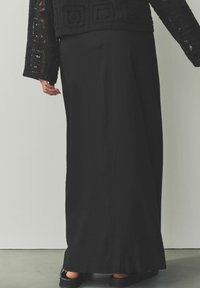 Falda maxi negra de tejido suave, con corte recto. Combina con un suéter de punto negro texturizado que presenta patrones abiertos.