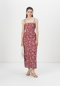 Mörkröd blommönstrad midiklänning med tunna axelband, figurnära överdel och utställd nederdel, kombinerad med en vit handväska och beige sandaler med remmar.