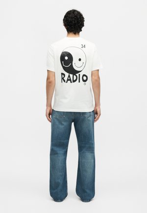 Άνδρας που φοράει λευκό T-shirt με ασπρόμαυρο σχέδιο yin-yang emoticon και το κείμενο «RADIO» στην πλάτη, συνδυασμένο με μπλε τζιν, στέκεται με την πλάτη προς το μέρος μας.