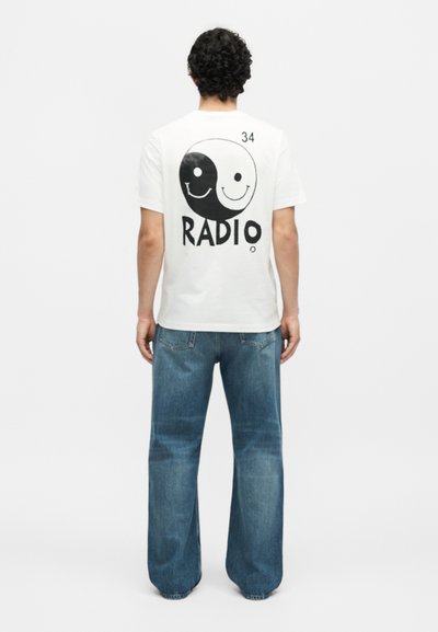 Άνδρας που φοράει λευκό T-shirt με ασπρόμαυρο σχέδιο yin-yang emoticon και το κείμενο «RADIO» στην πλάτη, συνδυασμένο με μπλε τζιν, στέκεται με την πλάτη προς το μέρος μας.