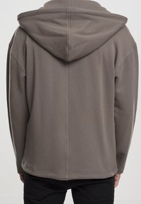 Persona che indossa un felpa grigia con cappuccio, vista da dietro, abbinata a pantaloni neri su uno sfondo semplice.