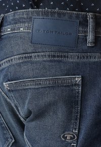 Jeans en denim bleu foncé avec une patch en cuir portant l'inscription "TOM TAILOR" à l'arrière. Comporte des poches arrière et des coutures contrastées.