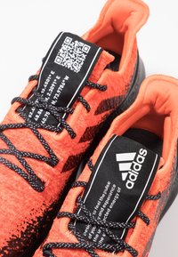 Jasně oranžové sportovní boty mají texturovaný svršek, černé detaily, výrazné tkaničky a štítek s QR kódem na jazyku.