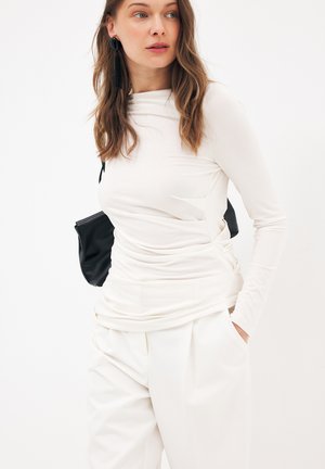 Donna che indossa una maglia bianca a maniche lunghe e pantaloni bianchi, con una pochette nera sotto il braccio, in piedi davanti a uno sfondo bianco.